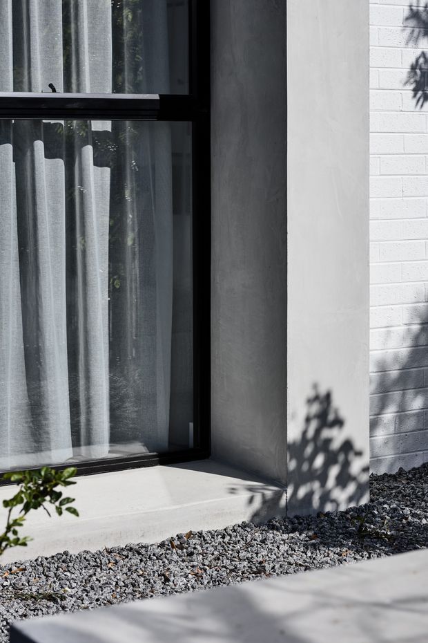 Close-up exterior architectural detail showing a large black-framed window with sheer curtains, a smooth light-grey rendered reveal, and adjacent white-painted brick wall. A concrete sill and gravel landscaping sit below, with strong sunlight and foliage shadows across the facade and paving.