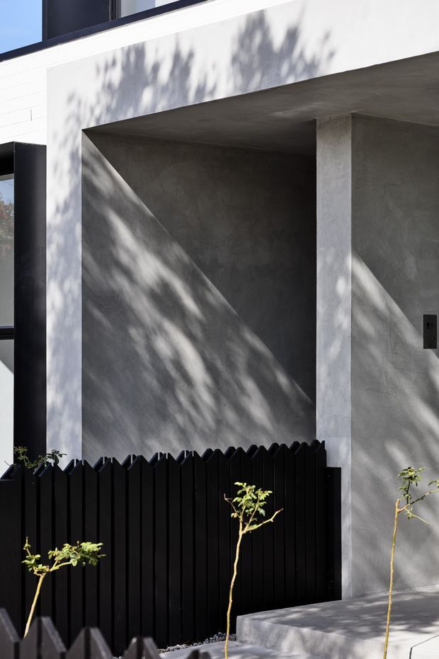 Close-up exterior view of a modern residential facade featuring a large recessed concrete entry or porch opening, smooth light-grey rendered/concrete surfaces, black-framed glazing, and a tall black vertical-slat fence. Strong dappled tree shadows fall across the facade and entry walls, with a simple concrete path and sparse planted trees in the foreground.