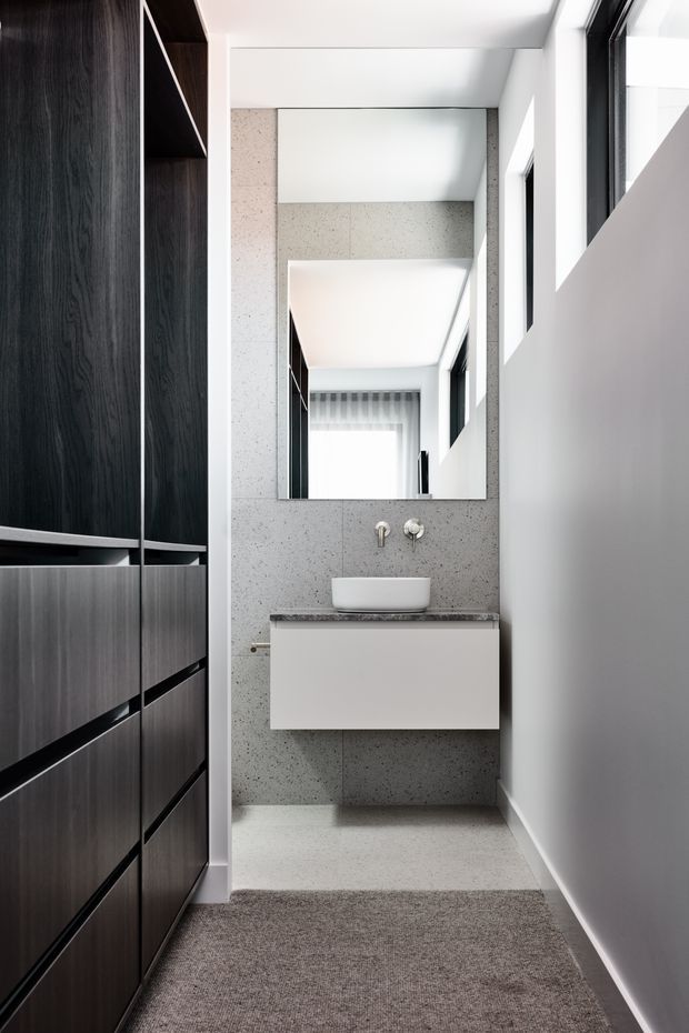 A narrow, contemporary bathroom or powder room with a wall-mounted white vanity, vessel basin, chrome fixtures and a large rectangular mirror. The back wall and floor are finished in light grey speckled tile or stone, contrasted with dark timber built-in cabinetry on the left and white walls. Black-framed windows and a soft grey carpeted approach add contrast and texture.