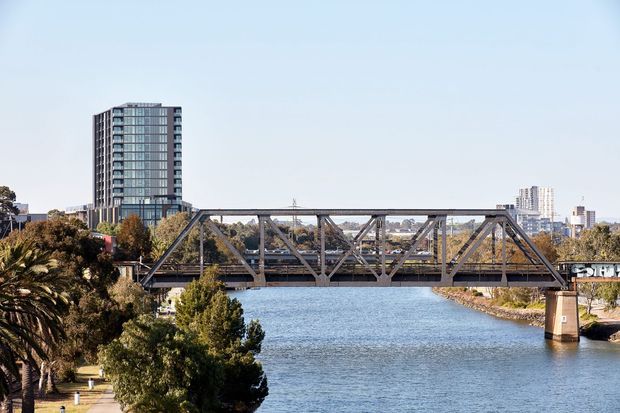 A daytime urban riverside view dominated by a steel truss bridge spanning a broad waterway. Trees and landscaped riverbanks frame the foreground, with modern high-rise buildings visible in the background beneath a clear blue sky.