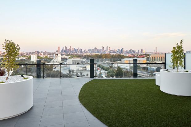A contemporary rooftop terrace or elevated outdoor space with large-format grey tiled paving, a curved area of green lawn or synthetic turf, white cylindrical raised planters with small trees, and a transparent glass balustrade with dark metal posts. The terrace overlooks a dense urban skyline in the distance under a bright, pale sky.