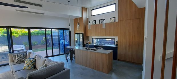 Open-plan contemporary kitchen and living area with polished concrete flooring, extensive warm timber cabinetry, a stone kitchen island with bar seating, integrated appliances, pendant lights, and large black-framed sliding glass doors opening to an outdoor area. A grey sectional sofa occupies the living zone, while high-level glazing brings in natural light.