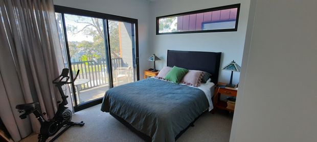 A contemporary bedroom with a double bed dressed in dark teal bedding, timber bedside tables, and black-framed glazing. A full-height glazed door opens to a balcony, while a narrow high-level window brings in additional light. An exercise bike sits near the curtained glazing, and the room has carpeted flooring and a neutral palette.