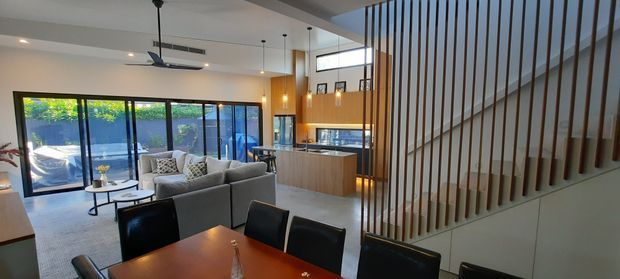 Open-plan contemporary home interior showing a dining area in the foreground, living room and kitchen beyond, and a timber-slat stair balustrade dividing the space. The room features warm timber cabinetry and dining furniture, light flooring, extensive glazing to the garden, pendant lights, and a high ceiling over the kitchen zone.