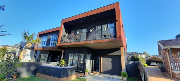 Exterior view of a contemporary two-storey residence with dark cladding, terracotta-toned upper facade framing, broad balcony, large glazed openings, integrated garage, and a small front garden. The home is situated among neighbouring suburban houses under a clear blue sky.