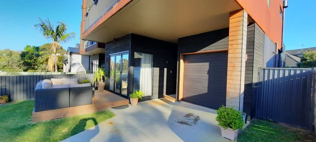 Contemporary multi-storey residential facade with a covered entry, dark cladding, warm timber-look vertical feature panels, large glazed doors, and an integrated single garage with a timber-toned roller door. A small landscaped lawn, outdoor seating area, driveway, and boundary fencing are visible.