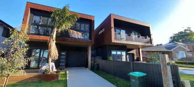 Street-facing view of a contemporary multi-level townhouse or residential duplex with dark charcoal cladding, warm reddish-brown vertical facade panels, large windows, projecting upper-level volumes, balconies, and a covered entry/carport area. A paved driveway, front garden planting, fencing, and neighbouring homes are visible.