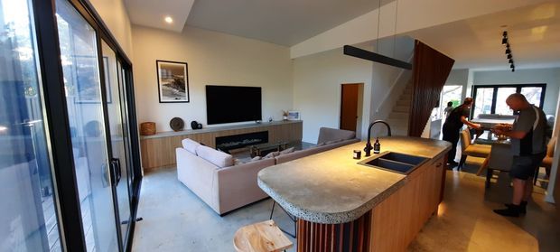 Open-plan contemporary kitchen and living area with a rounded stone island and integrated sink, timber cabinetry, pendant lighting, pale sofa, television, and a dining area beyond. Full-height glazing lines the left side, while a timber-clad stair feature adds visual warmth.