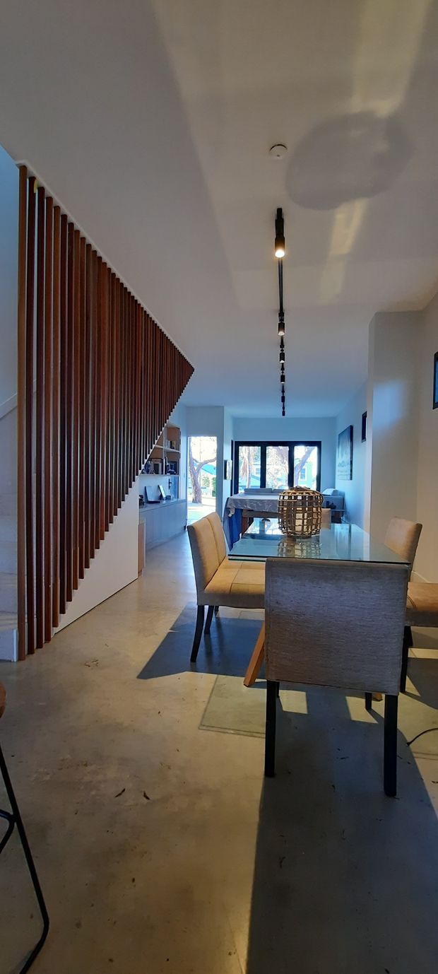 A contemporary open-plan dining and hallway interior viewed along a long, narrow space. A glass dining table with upholstered chairs occupies the foreground, while a sculptural timber vertical-slat screen conceals or borders the staircase on the left. Pale walls and polished concrete flooring are paired with a linear black track light, built-in cabinetry, artwork, and large glazed doors opening to the outdoors.