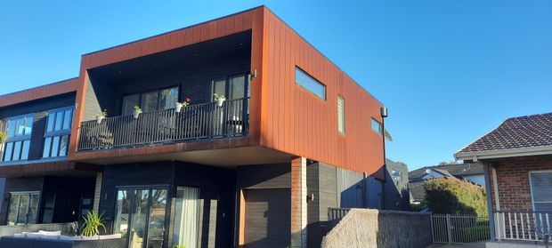 Exterior view of a contemporary multi-storey residential townhouse with a strongly projecting upper-level balcony, extensive dark-framed glazing, and warm rust-coloured vertical cladding. A neighbouring brick-roofed house is visible beside it under a clear blue sky.