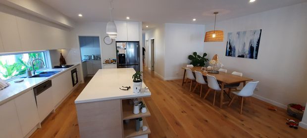 Open-plan kitchen and dining area with white flat-panel cabinetry, a large light stone island, stainless-steel appliances, warm timber flooring, and a timber dining table with contemporary white chairs. The space includes recessed ceiling lights, pendant lighting, indoor plants, and abstract wall art, creating a bright, warm, contemporary interior.