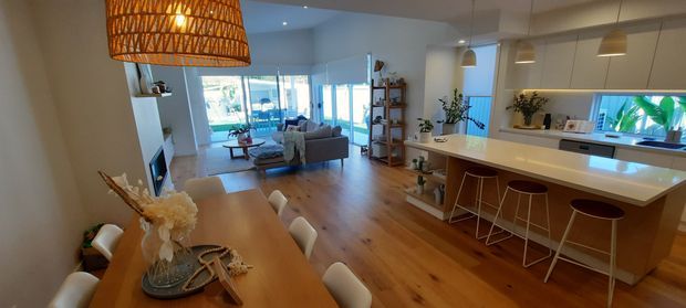 Open-plan residential interior combining a warm timber dining area, a contemporary white kitchen with island seating, and a living room opening to bright glazing. Natural woven pendant lighting, timber flooring, white cabinetry, and indoor plants create a relaxed, inviting atmosphere.