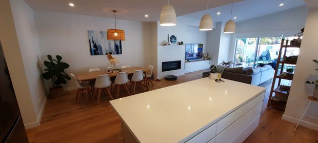 Wide-angle view of an open-plan contemporary kitchen, dining and living area. A large white island with integrated drawers dominates the foreground, with timber flooring, white cabinetry and walls, pendant lighting, a dining table, and a living area opening to a bright glazed outdoor space.