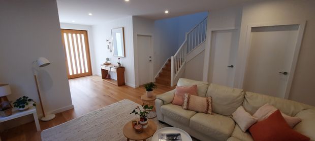 A bright, warm-toned open living area viewed toward an entryway and staircase. The room features a large cream sectional sofa with pink, beige and rust cushions, a pale textured rug, a round timber coffee table, light timber flooring, white walls and doors, recessed ceiling lights, and a timber entry door with vertical glazing. A staircase with white balustrade leads upward at the rear.