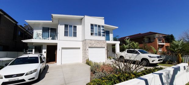 Street-facing exterior photograph of a contemporary multi-storey residential house with a white rendered and horizontal-clad facade, prominent pale stone feature wall, flat roof forms, balconies, and two integrated garage doors. The property sits in a suburban streetscape with landscaped planting and driveway parking.