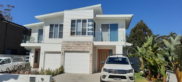 Front exterior view of a contemporary two-storey residence with a crisp white facade, asymmetrical low-pitched roof forms, projecting upper-level volumes, large windows, a stone-clad central garage surround, and an integrated ground-floor garage. Tropical planting and neighboring homes frame the property.