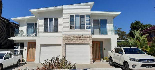 A bright daytime exterior view of a contemporary two-storey residential duplex or townhouse-style building. The facade combines white horizontal weatherboard cladding, white rendered sections, natural stone veneer around the central garage stack, timber front doors, large windows, glass balcony balustrades, and projecting low-pitched roof planes. Two garages and separate entries are visible, with neighbouring homes and vehicles in the foreground.