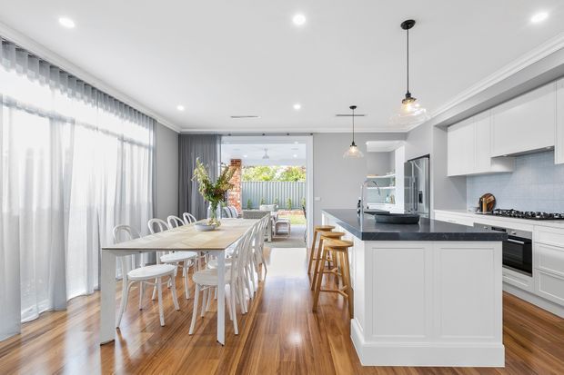 Bright open-plan kitchen and dining area with white shaker-style cabinetry, a large central island, timber flooring, and a long dining table beside full-height sheer curtains. Pendant lights, recessed lighting, and a glazed opening provide a clean, airy contemporary interior.