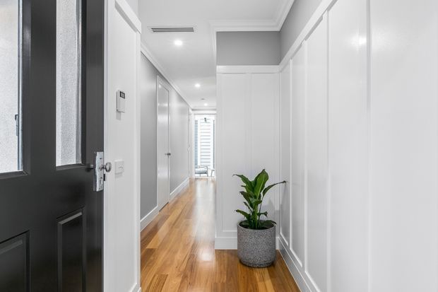 A bright, narrow residential hallway viewed from a black front door. The corridor features polished warm timber flooring, white paneled walls and built-in cabinetry, decorative ceiling cornices, recessed lighting, and a potted plant near the foreground. A glazed opening or window with horizontal blinds is visible at the far end.