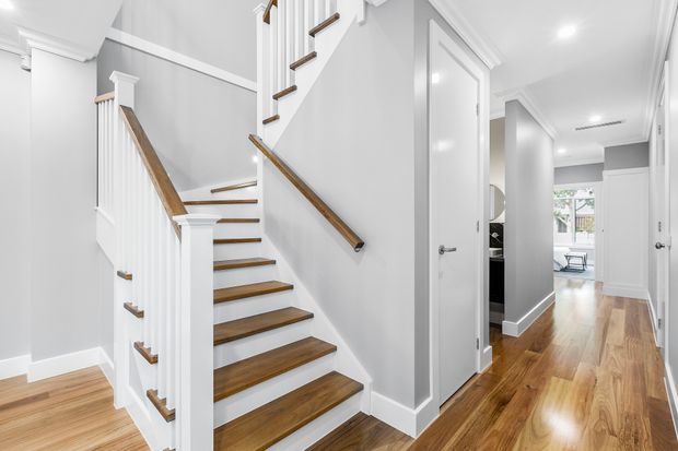 Bright, refined residential hallway with a central staircase featuring white painted balustrades, square newel posts, and timber handrails. Warm timber flooring continues through the corridor, with white trim, recessed ceiling lighting, and several white-panelled doorways leading to adjacent rooms.