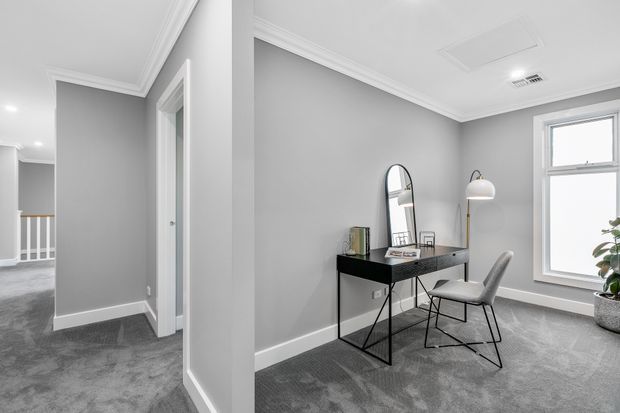 A refined home office occupies a grey-carpeted room with soft grey walls, white skirting boards and decorative cornices, and a large white-framed window. A slim black desk with geometric metal legs, an arched mirror, desk accessories, a white upholstered chair, and a dome floor lamp create a clean, contemporary workspace. A hallway and stair railing are visible through the opening to the left.