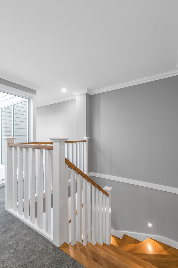 Bright residential stairway and landing with white painted balustrades, warm timber handrails and stair treads, grey painted walls, white crown moulding and trim, carpeted upper landing, and a large glazed opening to the side.