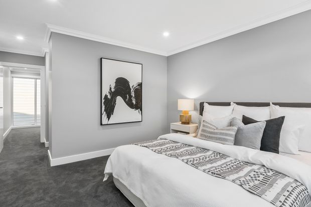A polished contemporary bedroom with a large upholstered bed dressed in white linens and layered grey and patterned cushions. The room has grey carpet, pale grey walls, white ceiling cornices and recessed lighting, with bedside tables and lamps. An open passage leads toward a bright adjoining area with window blinds.