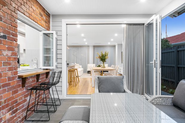 A covered outdoor entertaining area viewed toward a bright open-plan dining and living space through wide bi-fold doors. The terrace features a long outdoor dining table with upholstered seating, a built-in brick bar or serving counter with stools, and dark fencing. Inside, light timber flooring, white walls, grey curtains, and pale furnishings create a contemporary, airy interior.