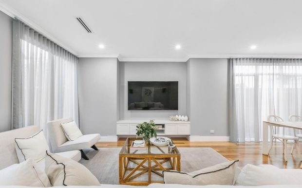 Bright, refined open-plan living and dining area with a neutral palette, sheer full-height curtains, pale timber flooring, a built-in television wall, white cabinetry, contemporary lounge seating, and a glass-topped timber coffee table.