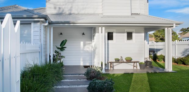 Bright exterior view of a white weatherboard home with a covered front porch, attached garage, small landscaped lawn, white picket fencing, and a grey metal roof. The facade features horizontal cladding, simple rectangular windows, and a sheltered entry with concrete stepping stones and garden planting.