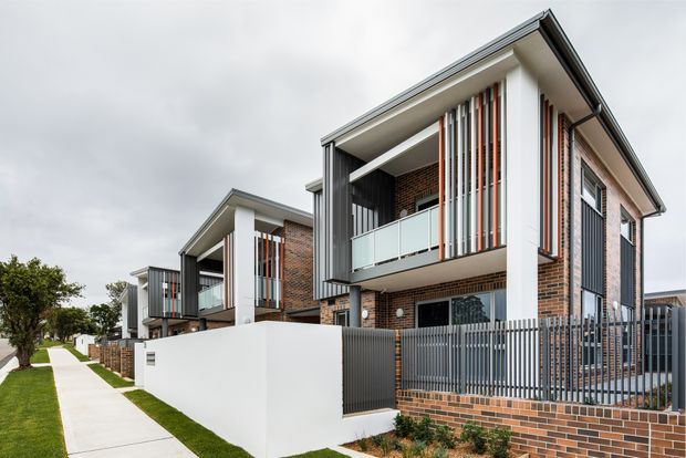 Street-facing view of a contemporary multi-storey townhouse development with repeated projecting balconies, exposed red-brown brick walls, white rendered structural framing, dark grey metal screening and fencing, and vertical timber-toned privacy battens. The homes sit along a landscaped pedestrian footpath beneath an overcast sky.