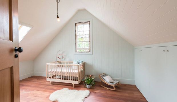 A bright, cozy attic nursery with a pitched sloping ceiling, pale vertical wall panelling, warm timber flooring, a wooden cot, rocking bassinet, built-in white cabinetry, and a small window. The room has a calm, softly styled character with natural daylight and minimal furnishings.