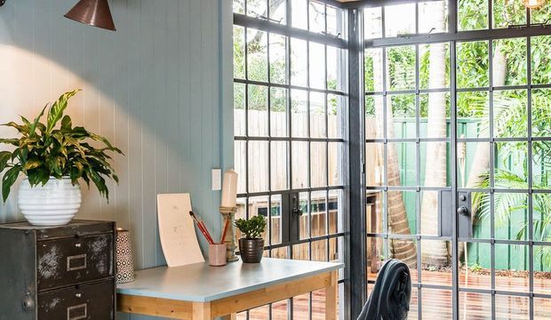 A bright, compact home office or study with a built-in-looking desk positioned beside large black-framed grid windows and glazed doors. The room has pale blue vertical wall panelling, a light blue-grey desk surface with timber edging, a dark vintage-style filing cabinet, copper pendant lighting, a potted plant, and a black office chair. Lush greenery and a timber deck are visible outside through the glazing.