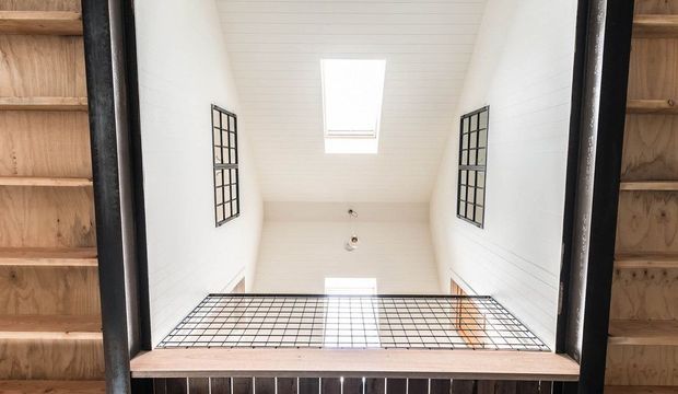 Looking upward through a dark-framed opening into a bright double-height interior void. White horizontal wall and ceiling panelling, a central skylight, high black-grid windows, exposed timber framing at the edges, and a black metal balustrade define the space.