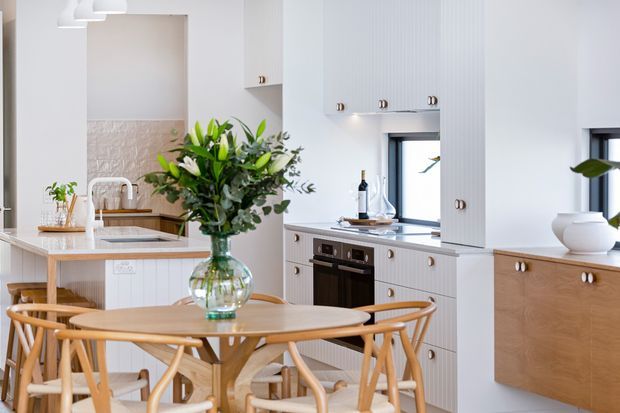 Bright, refined kitchen-dining interior with white vertically panelled cabinetry, light timber lower cabinetry, a stone-look benchtop, integrated oven, and a compact island. A round light-wood dining table with woven timber chairs occupies the foreground, while potted greenery and a glass vase add organic accents. Black-framed windows and warm timber details create a contemporary, welcoming atmosphere.