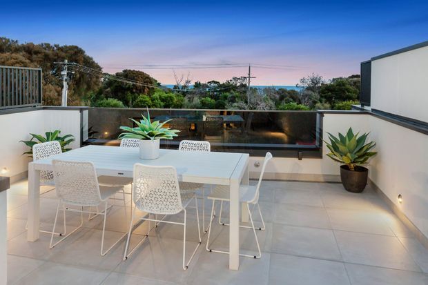 A modern rooftop or upper-level outdoor dining terrace with a white dining table and woven white chairs, overlooking a leafy coastal or suburban landscape. Pale tiled flooring, rendered privacy walls, potted plants, integrated wall lighting, and a dark glazed balustrade create a clean contemporary entertaining space at dusk.
