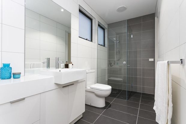Bright contemporary bathroom with white tiled walls, a wall-mounted white vanity and basin, bathtub, toilet, and a glass-enclosed shower. Dark grey floor and shower tiles create contrast, with a mirrored cabinet, recessed ceiling lighting, and a white towel rail.
