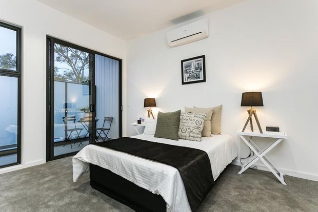 Bright, compact contemporary bedroom with a white upholstered bed dressed in white and black bedding, neutral cushions, bedside lamps, and a small white console table. Large black-framed glazing opens to a balcony with outdoor café furniture. Grey carpet, white walls, a wall-mounted air conditioner, and simple framed artwork create a clean, modern interior.
