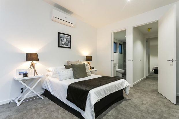 Bright, compact contemporary bedroom with a neatly made bed, layered white bedding, dark throw, bedside tables, table lamps, framed wall art, wall-mounted air-conditioning unit, and grey carpet. Open doors lead to an ensuite bathroom and an adjacent room or hallway.