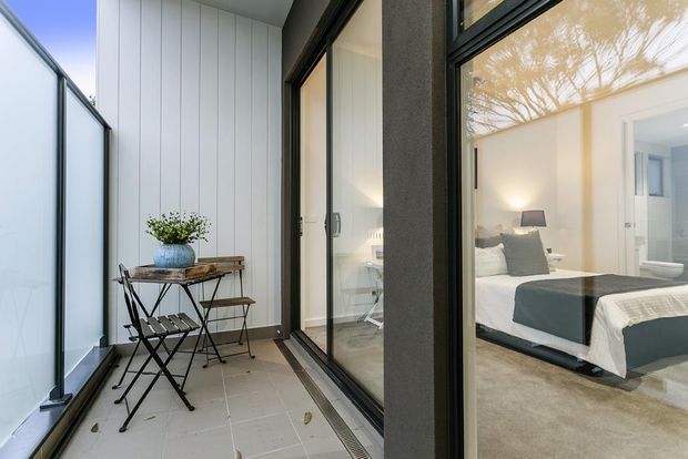 A narrow enclosed balcony or terrace with pale tiled flooring, white vertical wall panelling, and dark-framed glazing. A small bistro table and chairs sit beside the glass, overlooking a bright bedroom with a neatly made bed and neutral furnishings.