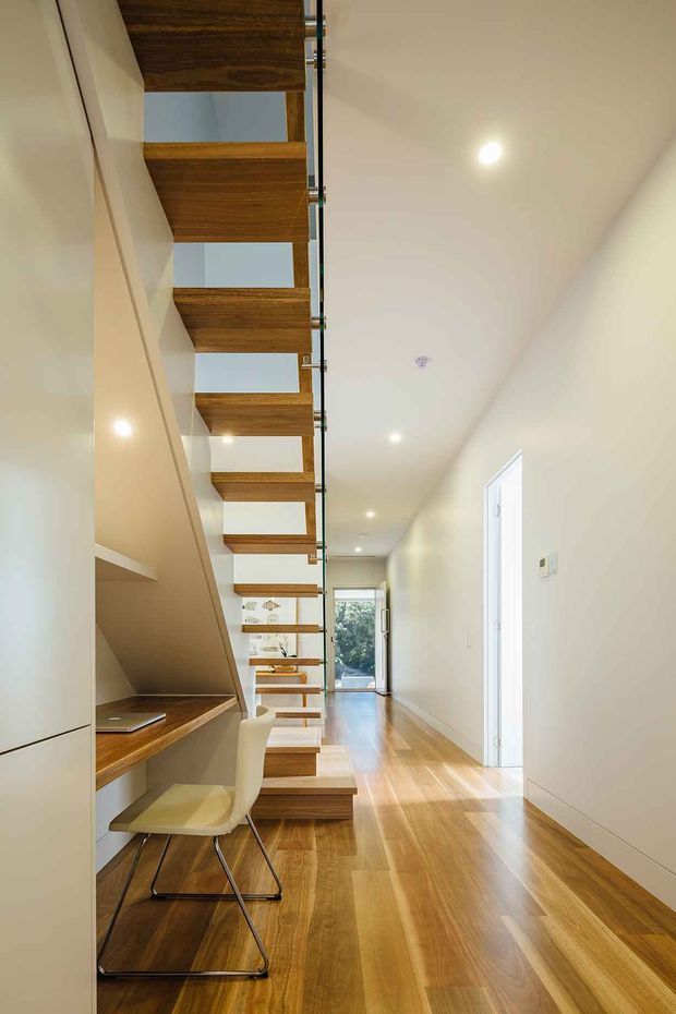 Bright, narrow contemporary hallway with warm timber flooring and a striking open-riser timber staircase. The stair structure incorporates integrated timber shelving, a compact built-in desk and chair beneath the stairs, with white walls, flush cabinetry and recessed ceiling lights. Glazing and a door at the far end bring in natural light.