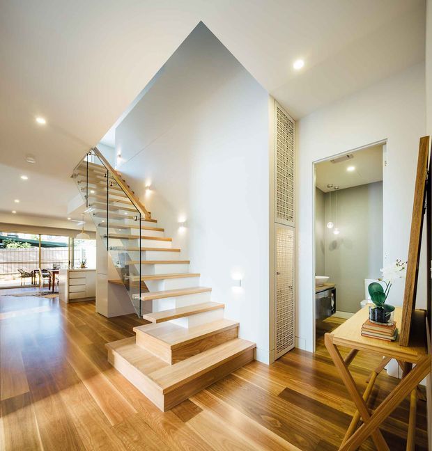 Bright contemporary interior view centred on a sculptural timber staircase with open risers, glass balustrades and a warm timber handrail. The staircase rises beside a tall white feature wall with a steep triangular upper profile. Warm timber flooring continues through the entry and dining area, while a bathroom is visible through an open doorway on the right. A narrow decorative screen and console table with mirror add detail to the entry zone.