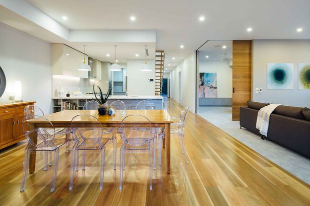 Open-plan contemporary interior combining a dining area, kitchen and living room. A long timber dining table with transparent acrylic chairs occupies the foreground, with timber cabinetry and a light kitchen behind it. The living area includes a dark sectional sofa, pale rug, abstract artwork and a timber feature wall with an open doorway. Polished timber flooring, white walls and recessed ceiling lighting create a bright, streamlined atmosphere.