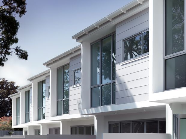 Exterior view of a contemporary multi-unit residential building with repeated white horizontal cladding, projecting upper-floor window bays, dark-framed glazing, and white roof gutters. The facade has a clean, rectilinear composition with sheltered ground-level entries or garage openings and low boundary walls. Mature trees and neighbouring roofs are visible in the background.