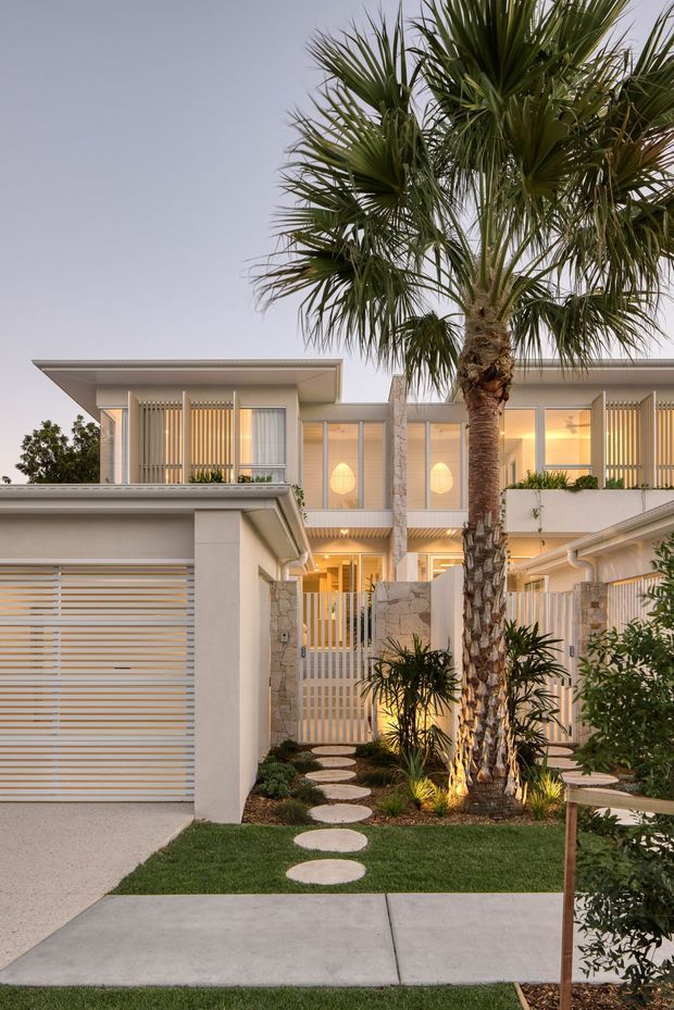 Contemporary multi-level residential facade viewed from the front, featuring pale rendered walls, broad flat rooflines, extensive glazing, vertical screening, and a white slatted garage door. A landscaped entry garden with a central palm, tropical planting, stepping stones, low stone walls, and warm architectural lighting leads toward the entrance.