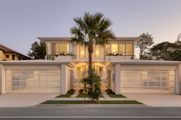 Symmetrical contemporary two-storey residential facade at dusk, with twin integrated garages, pale rendered walls, broad horizontal roof forms, screened upper-level glazing, a central gated entry, and a landscaped pedestrian approach with a palm tree. Warm interior lighting creates an inviting evening presentation.