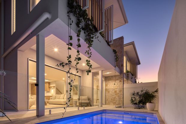 Twilight view of a contemporary multi-level residence centered on a small illuminated swimming pool and paved courtyard. The home features large glazed sliding doors, projecting upper-floor balconies with vertical timber screening, white rendered walls, a stone-clad feature wall, recessed downlights, and lush climbing and potted planting. Warm interior lighting contrasts with the cool blue pool and evening sky.