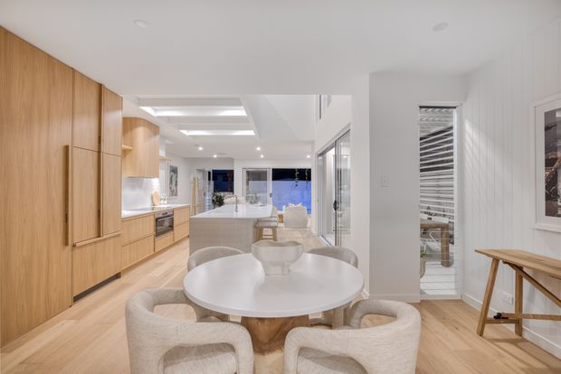 Bright open-plan contemporary interior combining a kitchen, dining area and living space. Full-height warm timber cabinetry lines the left wall, with a pale island and white stone-like benchtops at the centre. A round white dining table with upholstered curved chairs occupies the foreground, while white wall panelling, light timber flooring, recessed ceiling lighting and large glazed openings create an airy, refined feel.