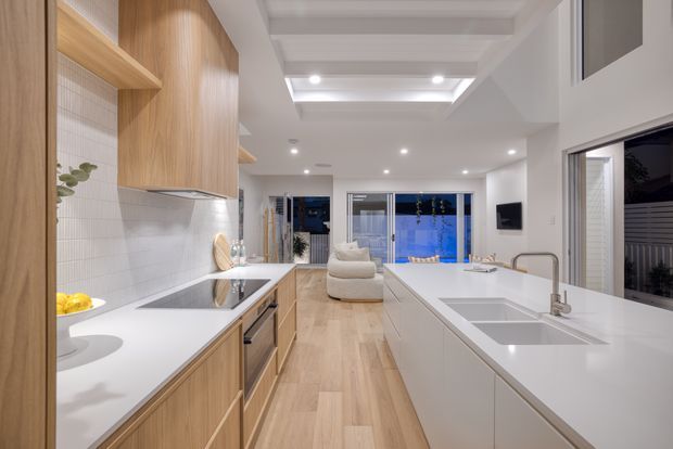 Bright open-plan kitchen and living area with a long white island, integrated double sink and brushed metal tapware, light timber cabinetry, white tiled splashback, and light timber flooring. The space opens toward a relaxed living area and large glazed doors, with a coffered ceiling and recessed lighting adding architectural definition.