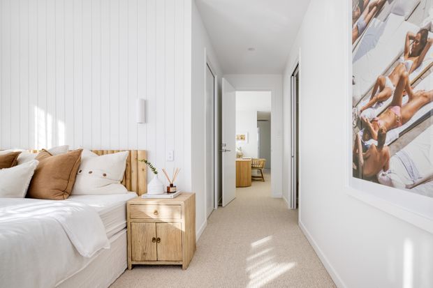 Bright, tidy bedroom opening onto a carpeted hallway. The room features a low bed with white bedding and tan cushions, a light timber slatted headboard, a matching timber bedside cabinet, white vertical wall panelling, and a large framed artwork. A bathroom or adjoining room is visible at the end of the hall.